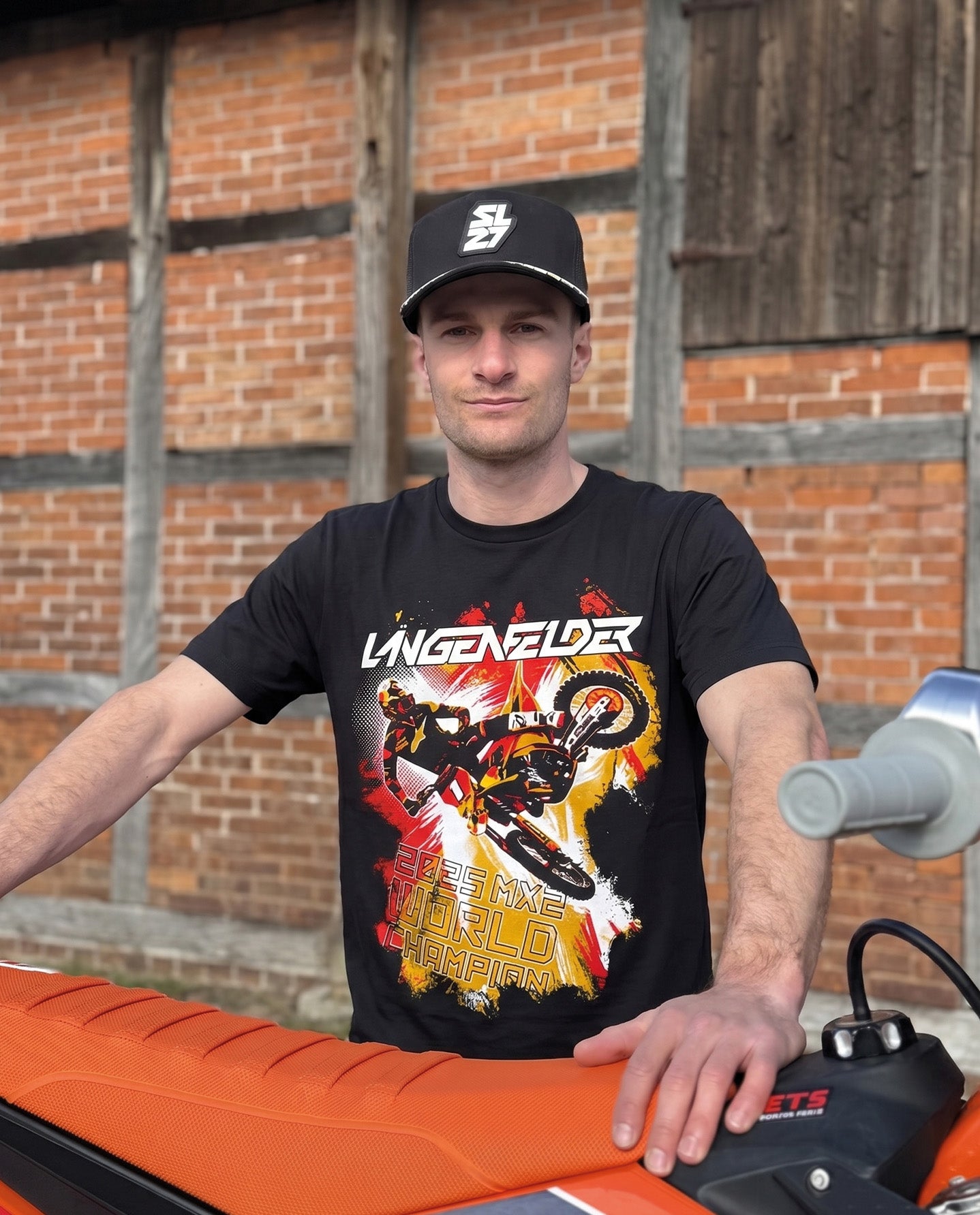 Mann mit schwarzem SL27 „HomeGP Germany“ T-Shirt steht neben einer KTM Motocross-Maschine. Das Frontdesign zeigt Simon Längenfelder in Racing-Action mit Deutschlandkarte in Schwarz-Rot-Gold und „2025 MX2 World Champion“-Schriftzug.