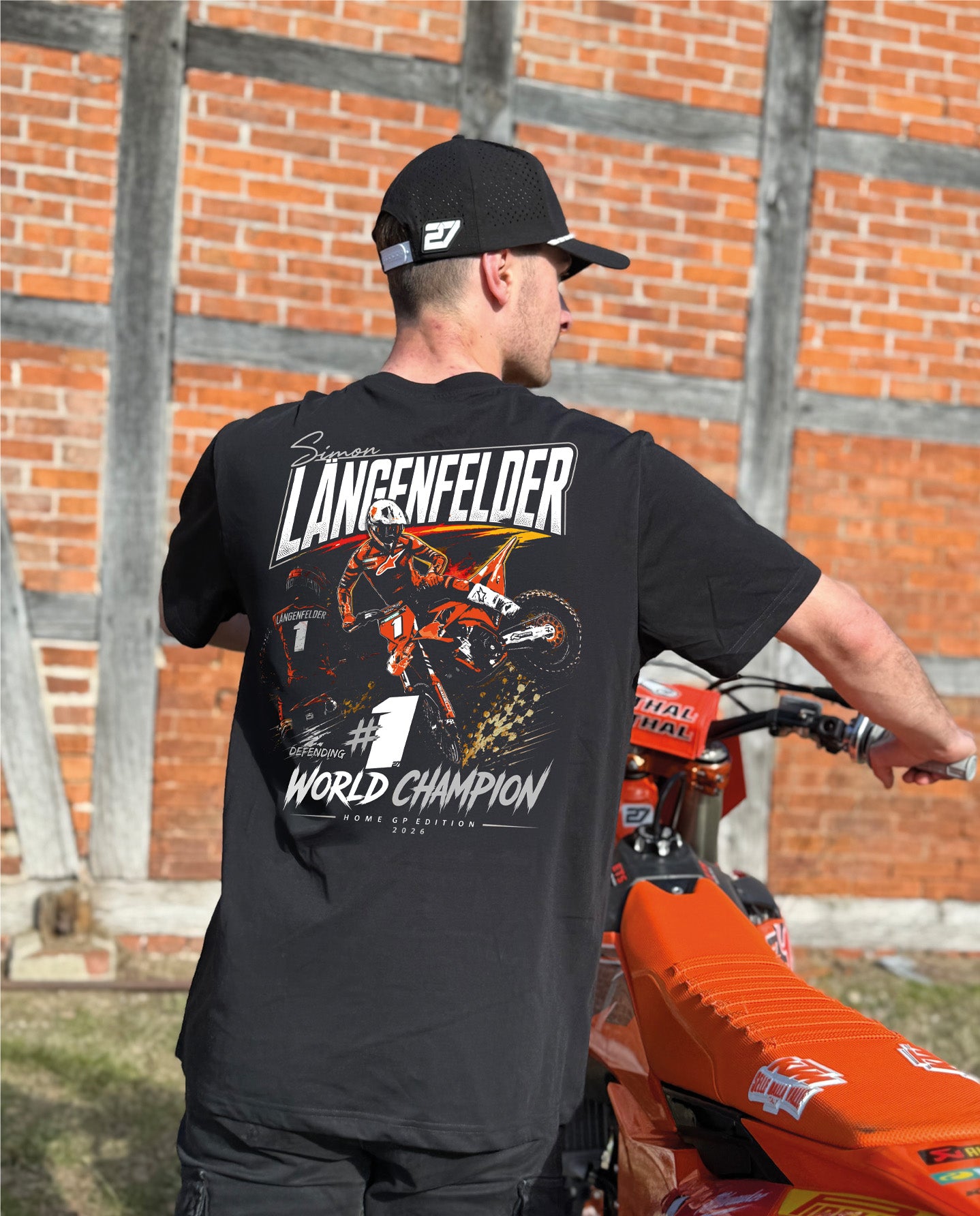Rückansicht eines schwarzen SL27 „HomeGP #1 World Champion 2026“ T-Shirts mit großem Rückenprint von Simon Längenfelder auf dem Motocross-Bike, kombiniert mit „#1 World Champion“-Schriftzug und Rennsport-Design in Orange, Weiß und Schwarz.