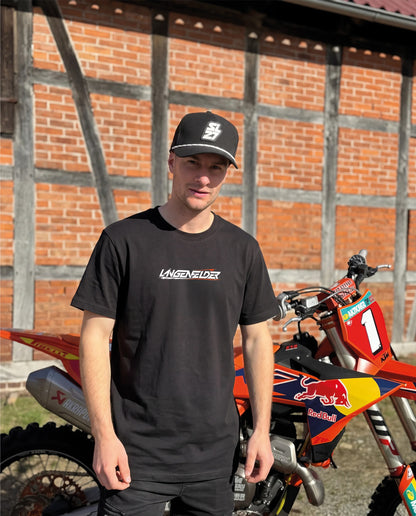 Mann mit schwarzem SL27 „HomeGP #1 World Champion 2026“ T-Shirt steht vor einer KTM Motocross-Maschine. Das Shirt zeigt vorne ein dezentes Langenfelder-Logo auf schwarzem Hintergrund.