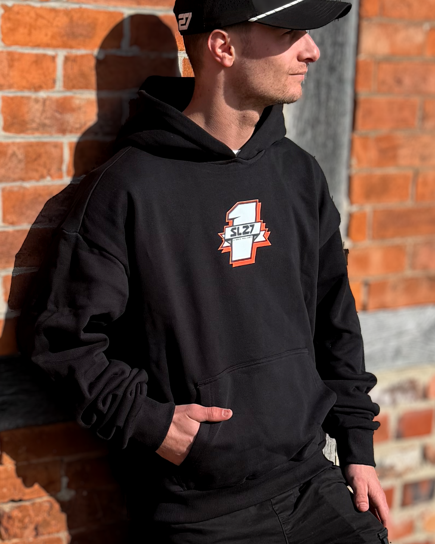 Schwarzer SL27 Hoodie „Number One“ mit kleinem Nummer-1-Frontlogo auf der Brust, getragen von Simon Längenfelder vor einer Backsteinwand.