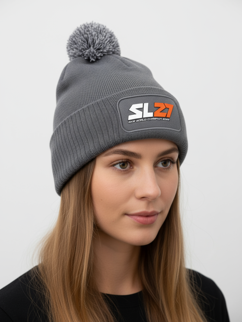 Graue SL27-Bommelmütze mit farblich passendem Pompon und schwarzem SL27-Gummipatch auf der Vorderseite, präsentiert vor hellem Hintergrund.