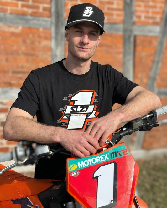 Schwarzes SL27 T-Shirt „Number One“ mit großem Nummer-1-Design und SL27-Logo auf der Vorderseite, getragen von Simon Längenfelder neben einem Motocross-Bike.