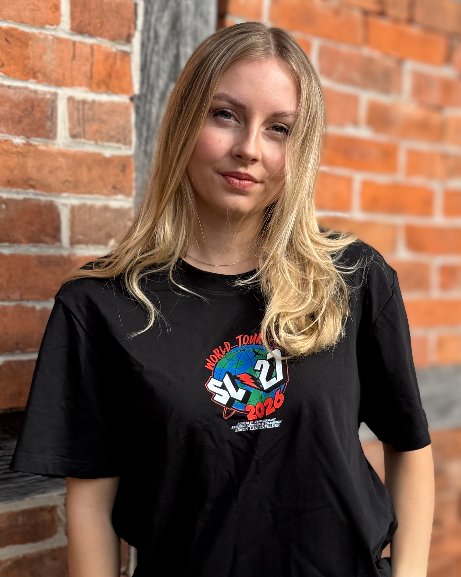 Schwarzes SL27 T-Shirt „World Tour 2026“ mit kleinem Frontlogo und Globus-Design, getragen vor einer Backsteinwand.