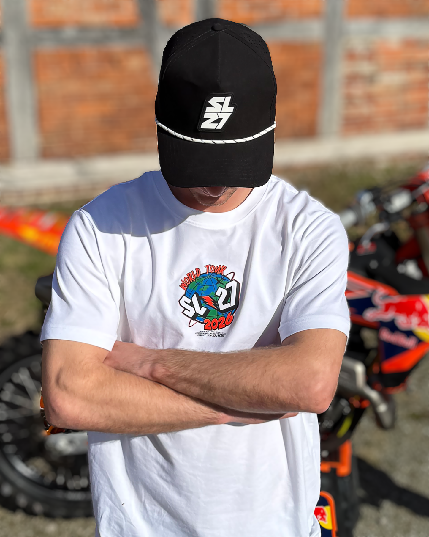 Weißes SL27 T-Shirt „World Tour 2026“ mit kleinem Globus-Frontlogo, getragen von Simon Längenfelder vor seinem Motocross-Bike.